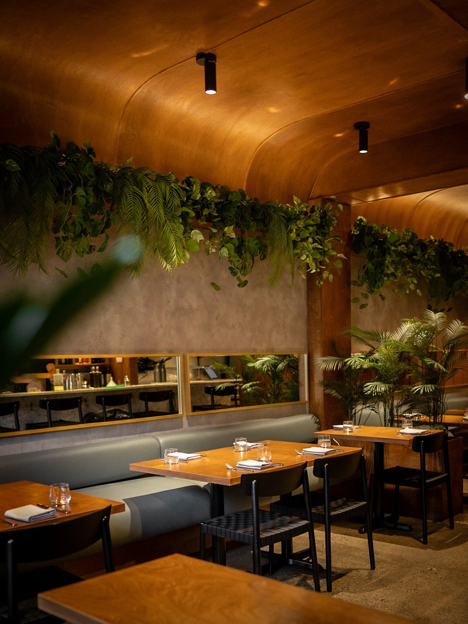 Een modern restaurant met houten tafels, zwarte stoelen, hanglampen en groene planten.